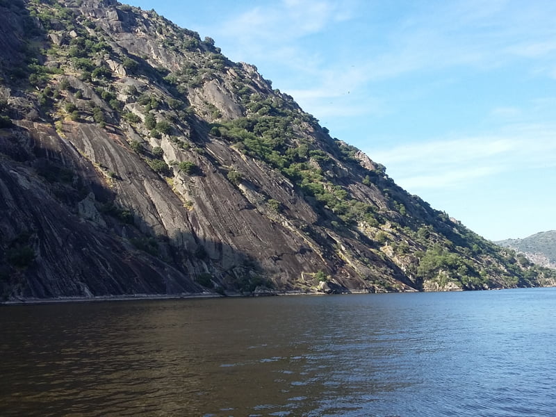 Steile rotswand naast kalme rivier onder blauwe hemel.