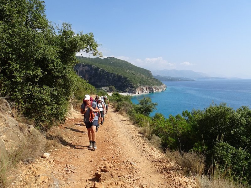 Wandelaars op ruig pad langs blauwe zee en heuvels.